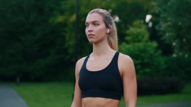 Parkta durmuş, kulaklık ve spor kıyafetleri giymiş kameraya bakan çekici kadın koşucunun portresi. İnsanlar ve spor konsepti