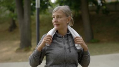 Smiled mature woman in sporty clothes carrying towel in her neck standing in the park after training. Portrait of elderly female looking at camera before the training
