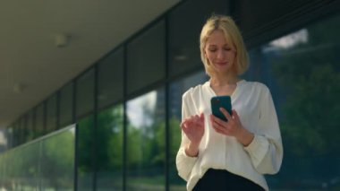 Ofisin arka planında duran resmi takım elbiseli çekici genç iş kadını cep telefonunu kullanıyor, müşterilerle mesajlaşıyor, uygulamaları çalıyor. İnsanlar ve teknoloji kavramı