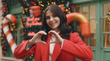 Portrait of beautiful caucasian woman feels happy and romantic shapes heart gesture standing on the winter decorated street. Love. Winter holidays. Valentines day