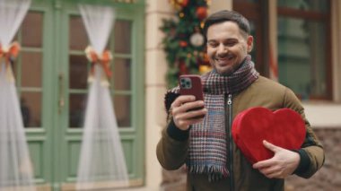 Handsome caucasian man standing in the winter decorating street holding heart shape gift and using smartphone. Handsome brunette outside, texting, scrolling apps online. Valentines day, online dating