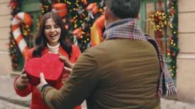 Happy couple in love. Valentines day. Romance, relationships. Husband gives his beautiful wife a red heart shaped gift. Lovey couple in love spending time together exchanging presents. Festive mood