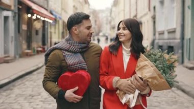 Happy cute couple walking in the winter city, talking together. Family in love going home, holding red heart shaped gift and christmas tree bouquet. Valentines day and people concept