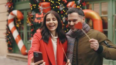 Happy caucasian couple doing selfie and holding the two paper hearts in their hands. Family standing in the winter decorated street, using smartphone, spending time together, feeling in love