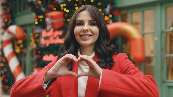 Portrait of beautiful caucasian woman feels happy and romantic shapes heart gesture standing on the winter decorated street. Love. Winter holidays. Valentines day