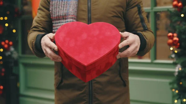 情人节 生日或结婚周年纪念日的红礼盒 由帅哥牵着 男子站在装饰过的冬日街道上 展示情人节礼物的特写 图库图片