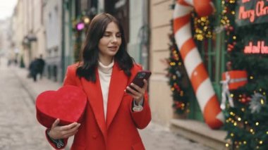 Happy woman walking in the decorating street holding the heart shape gift for valentine and using smartphone while snowing. Beautiful smiled brunette outside, texting, scrolling apps online