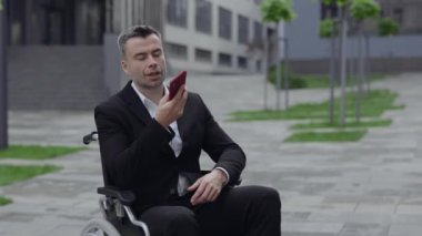Takım elbiseli başarılı bir adam caddede tekerlekli sandalyede otururken akıllı telefon kullanıyor. Engelli işadamı konuşuyor sesli mesaj yazdırırken, ses kaydı yaparken, dışarıda mikrofon kullanarak.