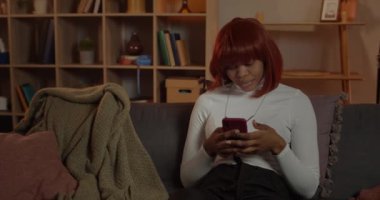 Genç ve çekici Afro-Amerikan kadın, evdeki kanepede otururken akıllı telefon ekranını kaydırıyor. Kızıl saçlı, gülümseyen lasy boş zamanlarında telefon kullanıyor.