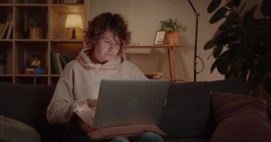 Woman Freelancer Using Laptop While Sitting on Couch at Home. Concentrated Female Person with Curly Hair Working Online Computer and Looking at Device Screen. Concept of Distance Work