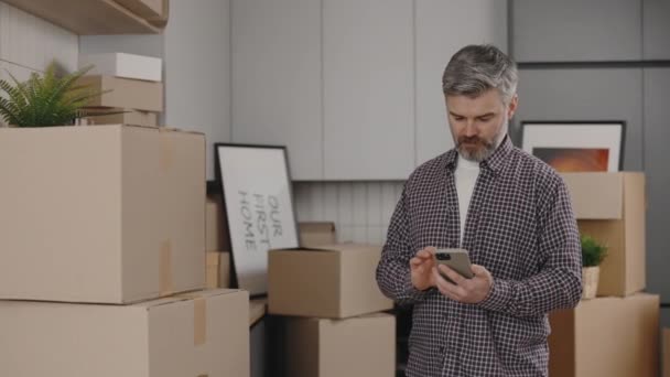 中年人在搬迁到新公寓时使用的是智能手机 新厨房背景上装有纸板箱 男子在流动电话上计算费用 — 图库视频影像