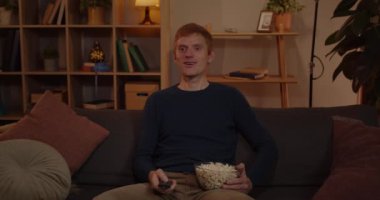 Kızıl Saçlı Adam Uzaktan Kumanda Kullanıyor ve Oturma Odası 'nda TV' yi açıyor. Koltukta rahatça oturup film izlerken patlamış mısırla kaseyi tutan heyecanlı genç adam.