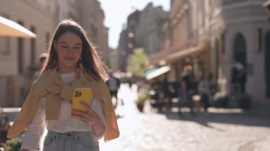 Şehir Caddesinde yürüyen ve akıllı telefon kullanan genç bir kadın. Açık hava güneşli. Cep telefonundan ekranda bilgi okuyan kız. Halk, Teknoloji ve İletişim Konsepti