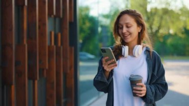Güzel, gülen beyaz kadın dışarıda akıllı telefon kullanıyor, mesaj atıyor, Eco Coffee kupasını tutuyor. Genç Bayan Çalışmak için Telefon Uygulaması Kullanıyor. Gadget ile Turist Seyahatte