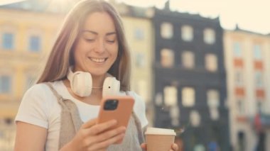 Gülümseyen Turist Kahve ve Ellerinde Dijital Cep Telefonu Kullanarak Sokağa Çıkıyor. Çekici Kafkas Mutlu Kadın Mesajlaşması, Kaydırma Uygulamaları. Teknoloji ve Yaşam Tarzları kavramı