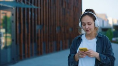 Güzel, gülümseyen beyaz bir kadın, akıllı telefon kullanıyor, büyük beyaz kulaklıklar takıyor, dışarıda yürüyor, mesaj yazıyor, not yazıyor ve müzik dinliyor. Halk ve Teknoloji Konsepti