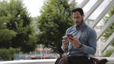 Açık havada bisikletin yanında oturan, kahve içen ve akıllı telefon kullanan resmi Afrikalı Amerikalı adam. Kulaklıklı İş İnsanının Çevrimiçi İletişimi Açık Hava.