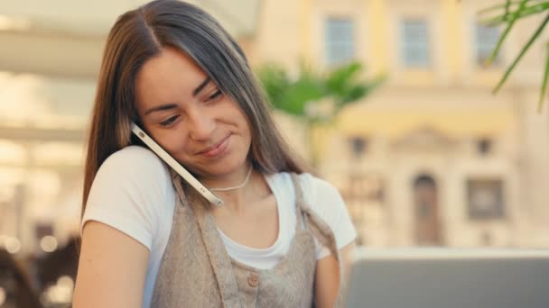 女性形象年轻白种人女孩坐在咖啡店外面看笔记本电脑屏幕在手机上说话友好电话 外面工作的可爱女人远程使用应用程序 — 图库视频影像