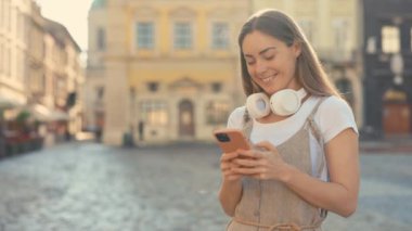 Cep Telefonu Kullanırken Şehirde Duran, Kablosuz Kulaklıklı Mutlu Genç Bayan Portresi. Mesajlaşma, internet taraması, sosyal medya. Akıllı Telefonla Yazan ve Kaydıran Şirin Kız