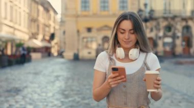 Şehir merkezinde yürürken iyi haberler okuyan çekici, beyaz bir kadın. Kahve ve Modern Akıllı Telefon 'la Sokaklarda Mutlu Turist Kız Yürüyüşü. Teknoloji ve Yaşam Tarzları kavramı