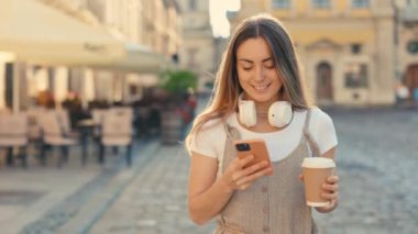 Cazip Kafkas Kadın Turist Şehir Sokakları boyunca yürüyor, elinde bir fincan kahve tutuyor ve Modern Akıllı Telefon kullanıyor. Mutlu kadın iyi bir şey okuyor ve gülümsüyor. Teknoloji ve Yaşam Tarzları Uzmanları