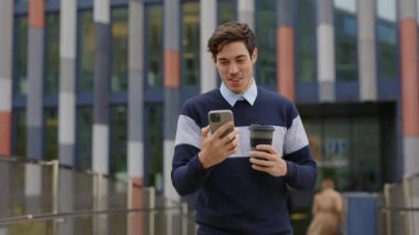 Yakışıklı genç iş adamı dışarıda yürüyor, cihazını kullanıyor ve iş konuşmalarına cevap veriyor. Patron İş Yeri 'ne yakın. Smartphone Holding Coffee' deki Uygulamalara Yazıyor ve Kaydırıyor.