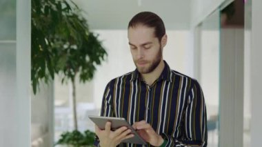 Koridorda dijital tablet kullanarak yürüyen uzun saçlı beyaz erkek ofis çalışanı. Cihaz üzerinde çalışan başarılı bir yönetici. Modern cihaz üzerinde çalışan genç bir serbest çalışan. İş ve Teknoloji