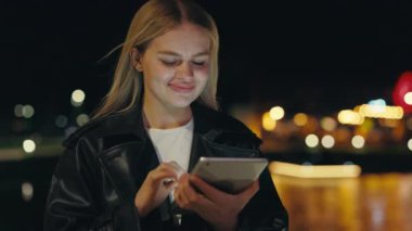 Gece Şehrinde Duran Gülümseyen Bayan Tablet 'e Sosyal Medya Yazıyor. Gülümseyen Mutlu Kadın internette geziniyor. İş Kadını Dışarıdaki Aygıtı Kullanıyor, Çevrimiçi Arkadaşlarla Sohbet Ediyor Mesaj Yazıyor