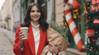 Beyaz kadın şenlik havasında kışın dekore edilmiş sokakta yürüyor, kahve içiyor ve elinde bir buket Noel ağacı tutuyor. Şehir merkezinde yürüyen esmer kadın. Kış tatili