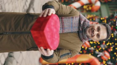 Dikey video. Beyaz sakallı adam kış süslü caddede elinde Valentino 'nun kalp şeklindeki hediyesiyle dikiliyor. Kırmızı Noel hediyeli yakışıklı adam dışarıda.
