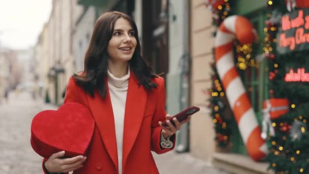 微笑的女人走在装饰街道上 手里拿着情人节的心形礼物 用智能手机 一边下雪一边环顾四周 外面漂亮的黑发 发短信 滚动应用程序 — 图库视频影像