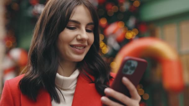 白种人笑着站在圣诞装饰街上用智能手机 外面漂亮的黑发 发短信 在线滚动应用程序 冬天的时候人与技术概念 — 图库视频影像