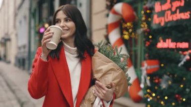 Beyaz kadın şenlik havasında kışın dekore edilmiş sokakta yürüyor, kahve ya da çay içiyor ve bir buket Noel ağacı içiyor. Şehir merkezinde yürüyen esmer kadın. Kış tatili