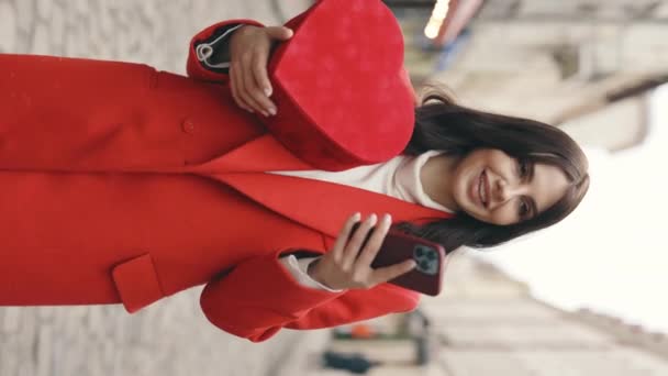 バーティカルビデオ 冬の通りを歩く白人女性は バレンタインの心臓形の贈り物を保持し 雪が降っている間スマートフォンを使用しています 外で美しいブルネット テキスト オンラインでアプリをスクロール — ストック動画