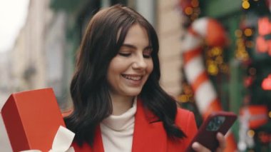 Şen şakrak bir kadın kışın dekorasyon sokağında elinde kırmızı bir hediyeyle yürüyüp akıllı telefon kullanıyor ve kar yağarken etrafa bakıyor. Dışarıda gülümseyen güzel esmer, mesaj atıyor.