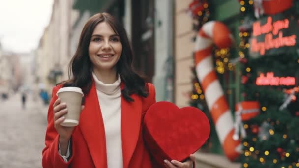 冬に装飾された通りを歩くお祝いの気分の白人女性 コーヒーや紅茶を飲んで 心臓の形のバレンタインギフトを保持しています 赤いプレゼントで市内中心部を歩いているブルネット女性 — ストック動画