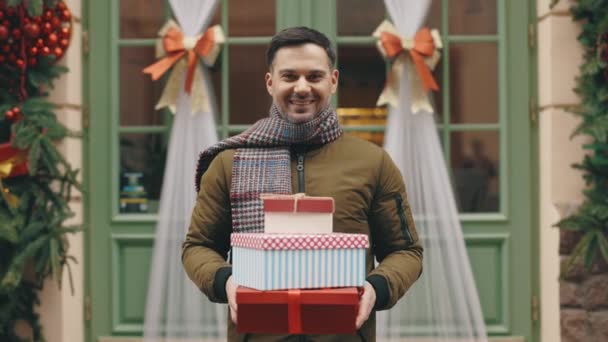 一个快乐英俊的男人站在一座装饰过的建筑物的背景上 并持有新年礼物的画像 带着积极胡子的家伙带着节日礼物向摄像机展示了他们 — 图库视频影像