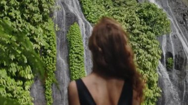 Sıska Kıvırcık Kız Ormanın Dışında Duruyor ve Şelalenin tadını çıkarıyor. Siyah Giysili Şirin Genç Bayan Doğaya Bakıyor. Rahatlama ve Turizm Konsepti