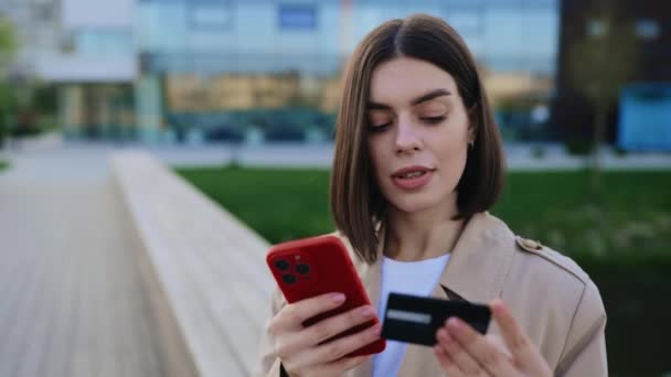迷人的年轻女士在外面使用智能手机和银行卡 参与网上移动购物 快乐女孩 在网上购物或购物时 坐在公园内 — 图库视频影像