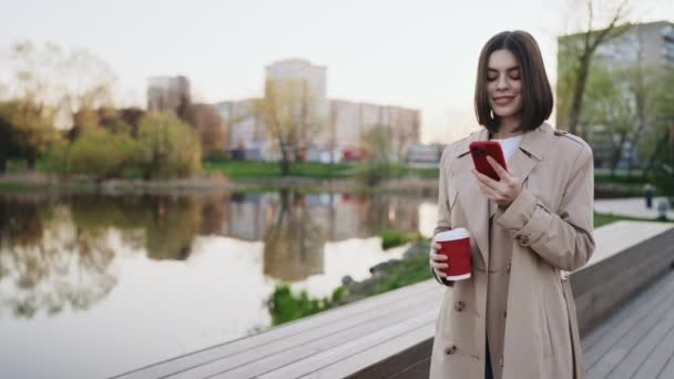 可爱的女人穿着丁字裤在当地湖畔散步 用智能手机 拿着咖啡杯 女孩在看智能手机屏风举行茶 网上购物 社交网络概念 — 图库视频影像