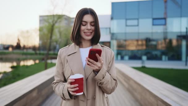 优雅的女人走在当地的湖边 一边手拿着咖啡一边用手机聊天 迷人的女孩一边上网一边去工作的地方喝咖啡休息 人与技术概念 — 图库视频影像