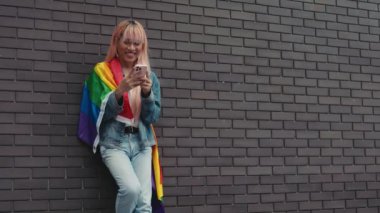 Makyajlı Yakışıklı Adam Şehir Merkezi 'nin Duvarında Duruyor ve Arkadaşlarıyla Sohbet Ediyor. Dışarıda LGBT bayrağı taşıyan olumlu adam Smartphone kullanıyor. LGBT ve Teknoloji Konsepti