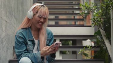 Pembe Peruklu Çekici Genç Adam Oturuyor ve Kulaklıkla Müzikten Hoşlanıyor, Cep Telefonuyla Sohbet Ediyor. Smartphone Kullanarak Sosyal Medya 'da Transseksüel Erkek Cevap Veriyor. LGBT ve Teknoloji Konsepti