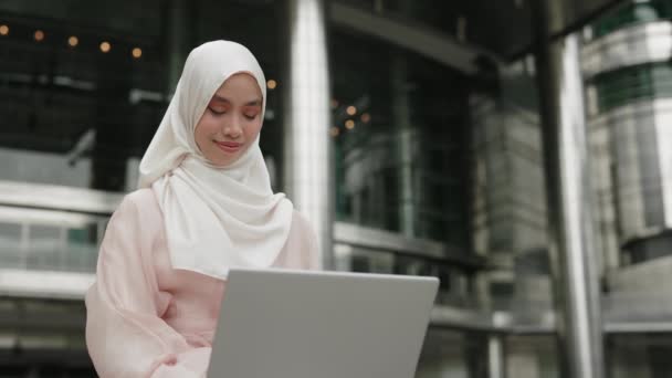 ヒジャブを着用している若い女性はオフィスビル外に座り ラップトップでリモートで作業します チャーミングでポジティブなイスラム少女の学生は屋外で学び 記事またはエッセイを書いています 人と技術のコンセプト — ストック動画