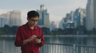 Gülümseyen yaşlı adam gölün yanında yürüyor ve Smartphone 'da sosyal medyada aşağı kayıyor. Üst düzey erkek açık hava cep telefonu kullanıyor, internette geziniyor. Halk ve Teknoloji Konsepti