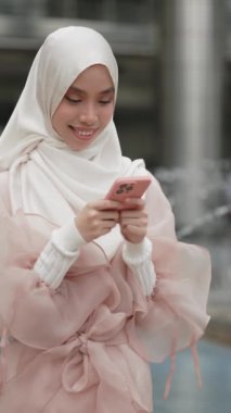 Beyaz tesettürlü, pembe elbiseli, akıllı telefonuyla sohbet eden sevimli Müslüman kız. Modern şehirde akıllı telefon kullanan Asyalı bir kadın. Dikey video
