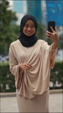 Dikey video. Dışarıda Selfie Yapan Çekici Kız telefonda ve dışarıda başörtüsü takıyor. Müslüman Kadın, cep telefonuyla sokakta resim çekiyor. Halk ve Teknoloji
