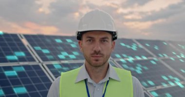 Yakışıklı beyaz erkek mühendis bir güneş enerjisi çiftliğinin önünde kameraya bakıyor. VFX grafikleri arka planda küresel ağ oluşturma ve yenilenebilir enerji teknolojisi kavramlarını canlandırır.