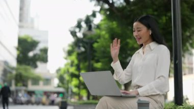 Neşeli Asyalı Kadın dizüstü bilgisayarıyla dışarıda oturuyor, Video Çağrısında Ekranda Selamlıyor. Mutlu İş Kadınları Video Konferansında Çalışmak İçin Bilgisayar Kullanıyor, Webinar 'a Katılıyor