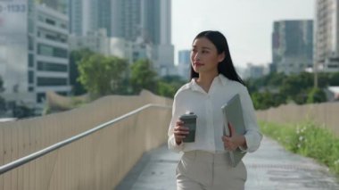 İşe gitmeden önce şehirde dolaşan, müşterileri için evrakları taşıyan inanılmaz genç bir koç. Mutlu Asyalı Kadın Kahve ve Önemli Belgeler Tutuyor, İş Görüşmesine Yürüyor